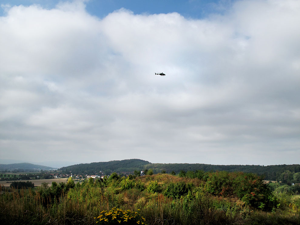 A Performer is flying in a Helicopter Bell 407 and looking at the exhibition from above for 2-3 minutes and then flying away.